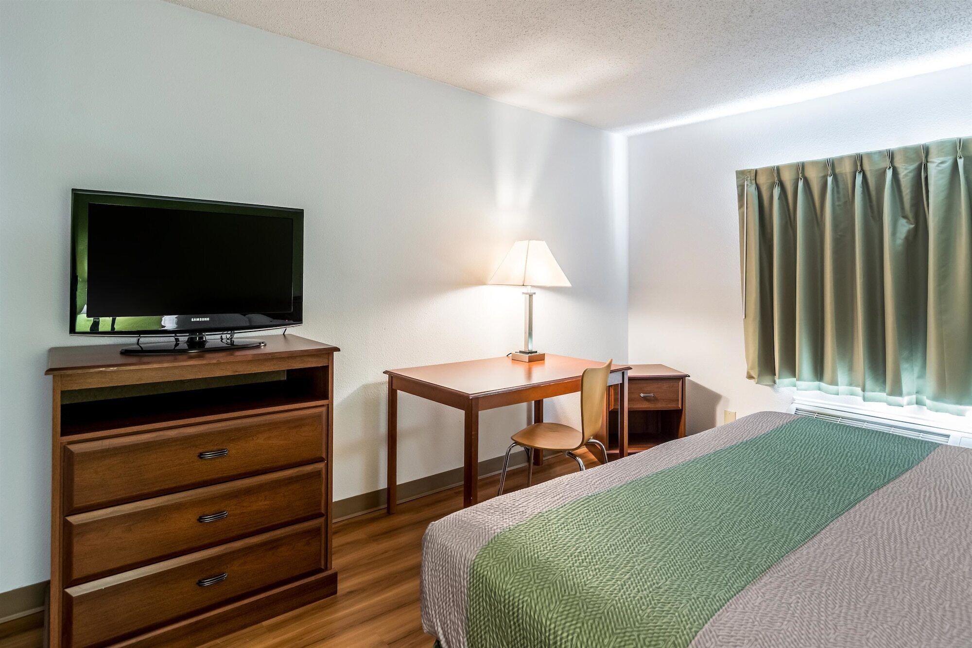 Une chambre d’hôtel comprenant un lit, un bureau avec une chaise, une télévision, une lampe et une fenêtre avec des rideaux.