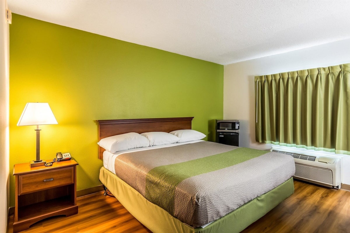 Habitación de hotel con cama, mesita de noche con lámpara, teléfono, una pequeña mesa, calefactor y ventana con cortinas.