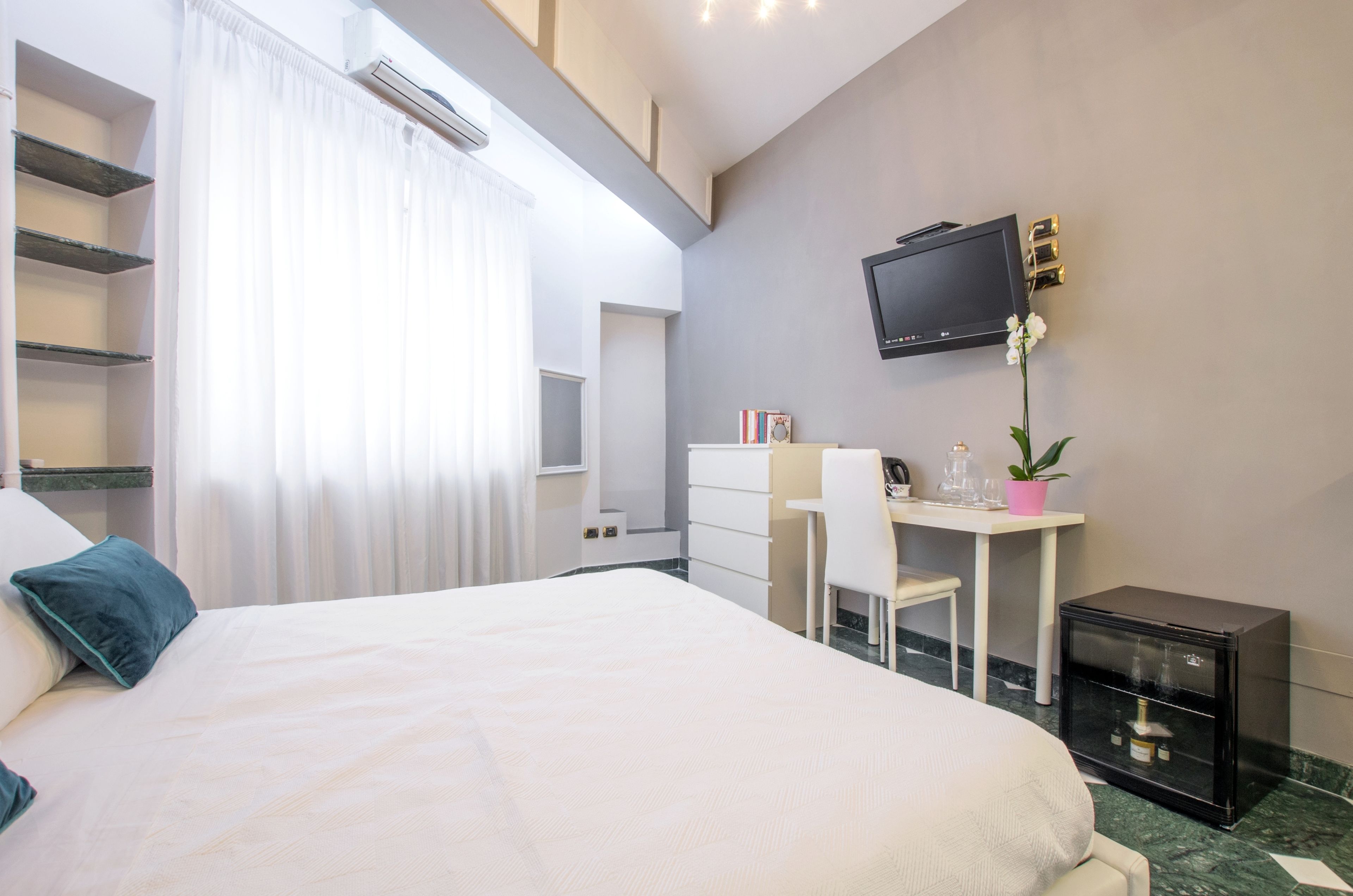A hotel room with a bed, a desk with a chair, a television, a small cabinet, and a potted plant.