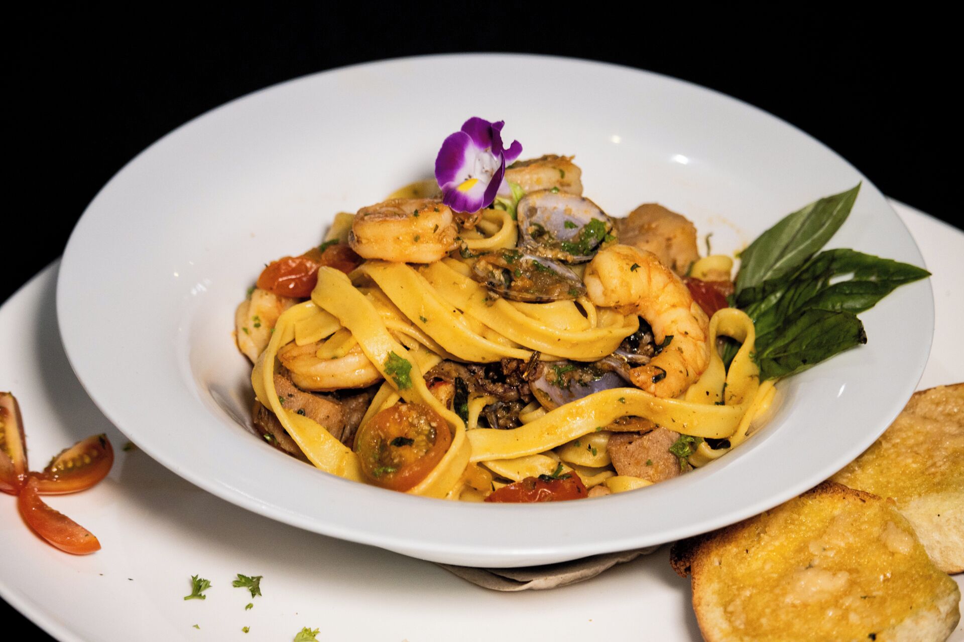 Ein Teller Pasta mit Garnelen, Muscheln und Gemüse, verziert mit einer violetten Blume und Basilikumblättern.