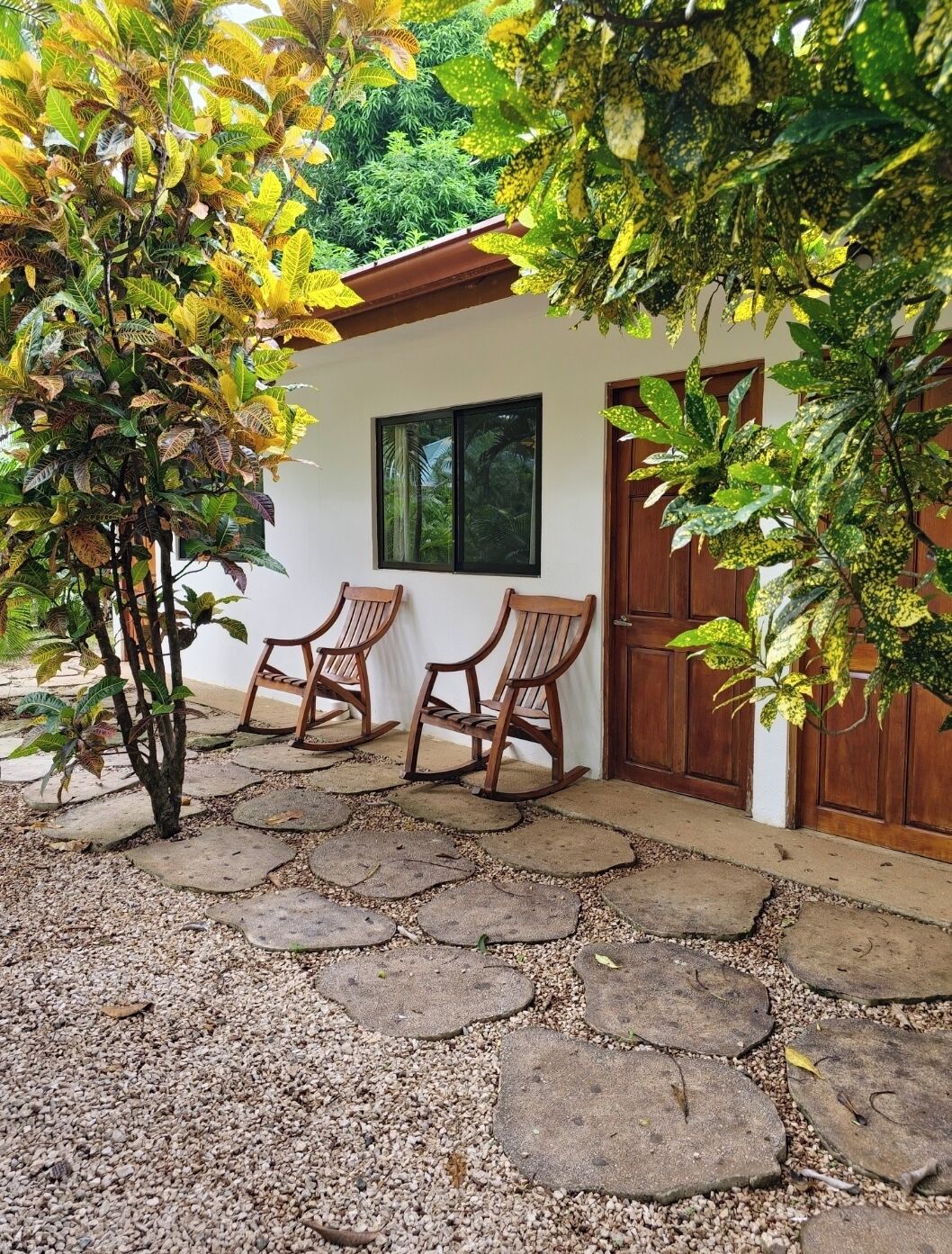 A patio with two wooden rocking chairs, a white building with a wooden door, and a window overlooking greenery.