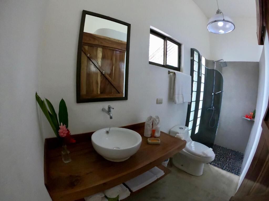 Un baño con tocador de madera, un lavabo blanco, un espejo, toallero, ducha y una pequeña ventana.