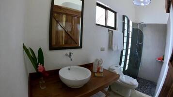 Un baño con tocador de madera, un lavabo blanco, un espejo, toallero, ducha y una pequeña ventana.