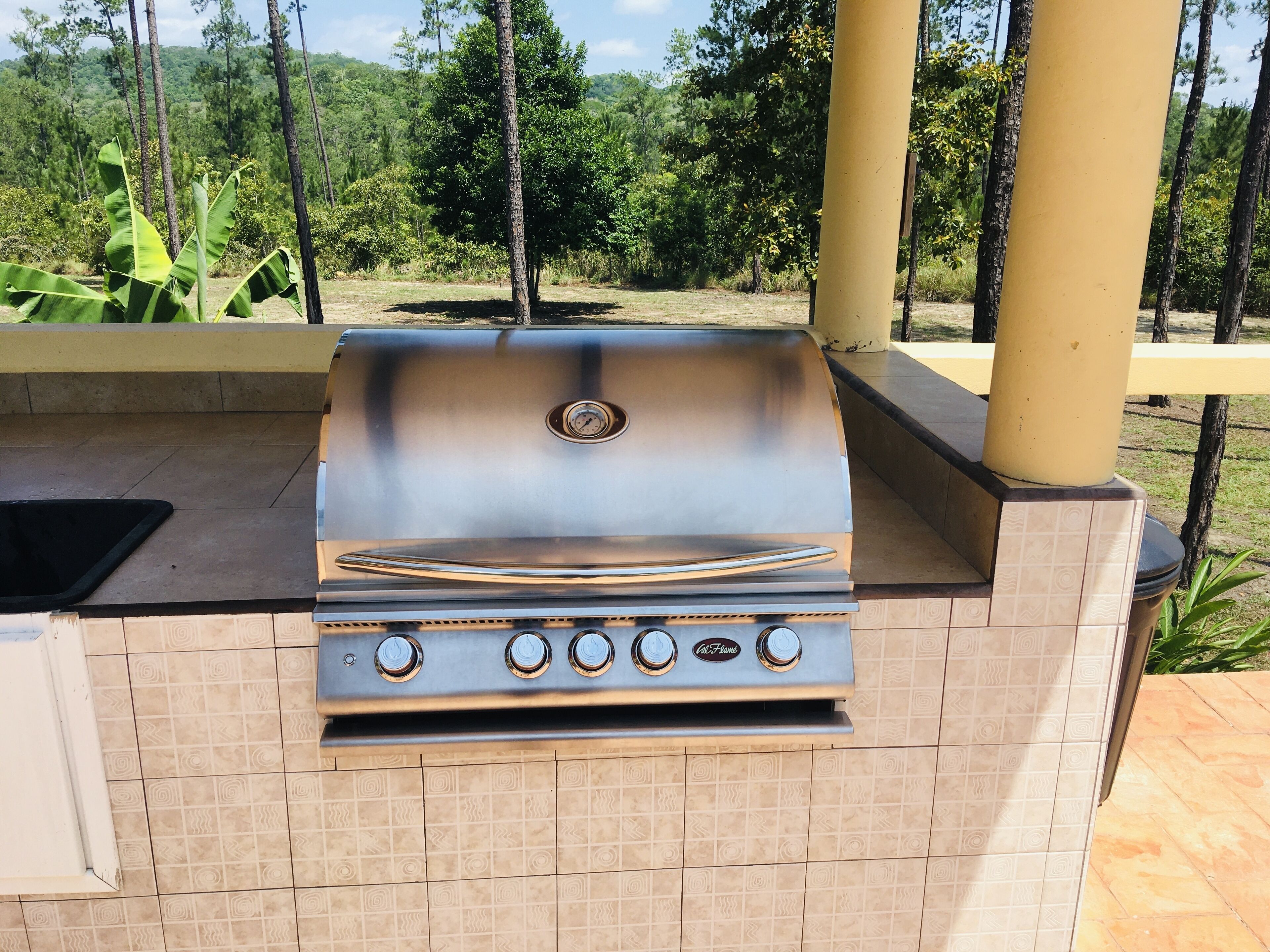 An outdoor grill with four burners and a stainless steel lid, set against a tiled wall with a view of a forested area.