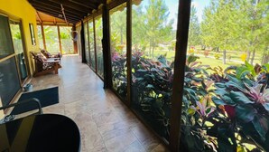 Un espacio al aire libre cubierto con una larga mesa y sillas, rodeado de grandes ventanales que dan a un jardín con diversas plantas y árboles.