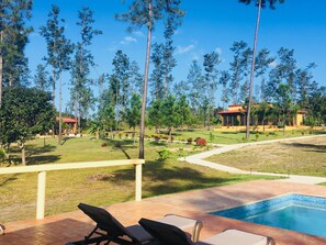 A poolside area with lounge chairs, a clear view of a landscaped garden, and a building with a red roof in the background. - Kāne Villas (Mountain Pine Ridge)
