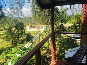A wooden balcony with a view of a lush garden, a pool, and a distant building. - Kāne Villas (Mountain Pine Ridge)