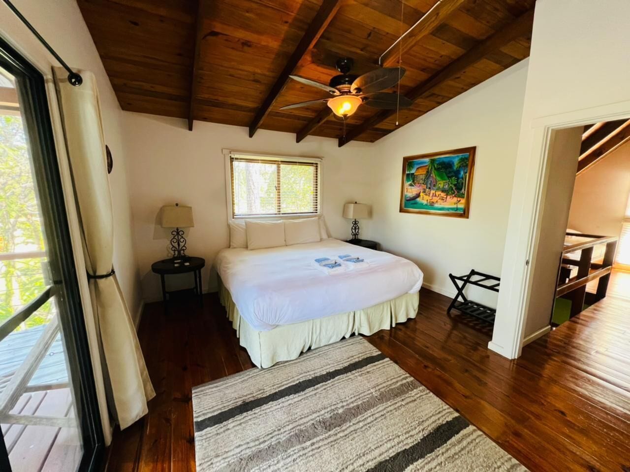 A bedroom with a wooden ceiling, a bed with a white bedspread, a window with blinds, a painting on the wall, and a wooden floor.