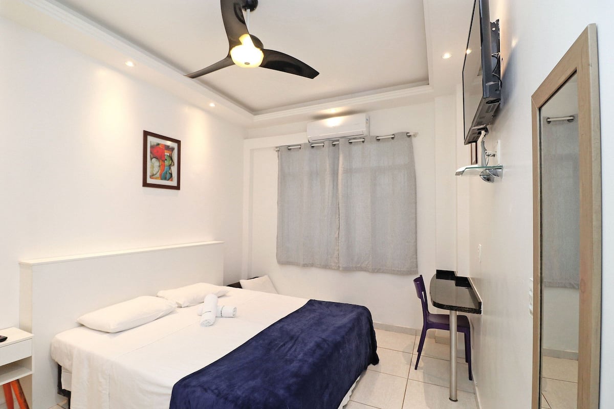 A bedroom with a bed, a desk, a chair, a ceiling fan, and an air conditioning unit.