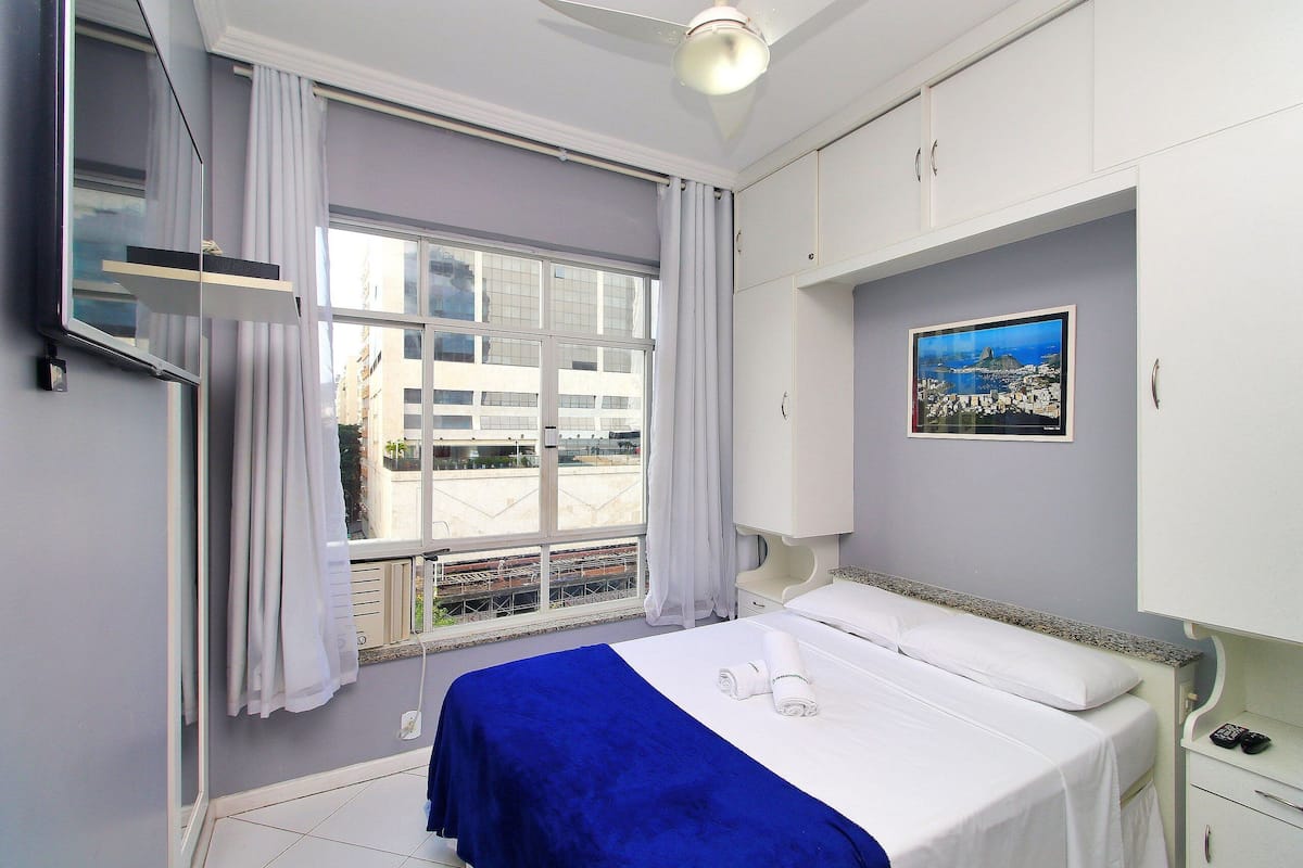 A hotel room with a bed, a window with curtains, a bedside table, and a wall-mounted TV.
