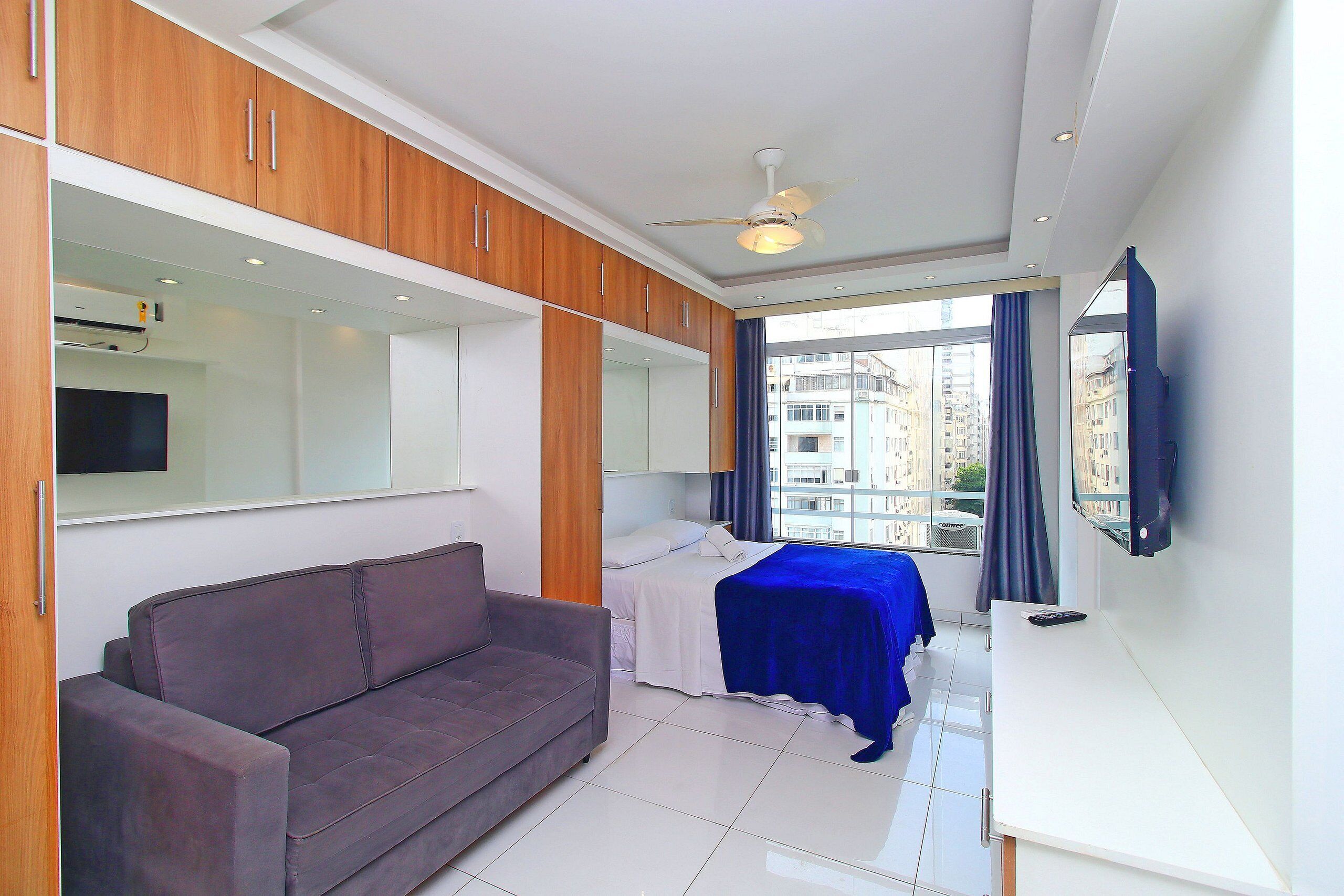 Quarto de hotel moderno com sofá, cama com colcha azul, escrivaninha e ventilador de teto.
