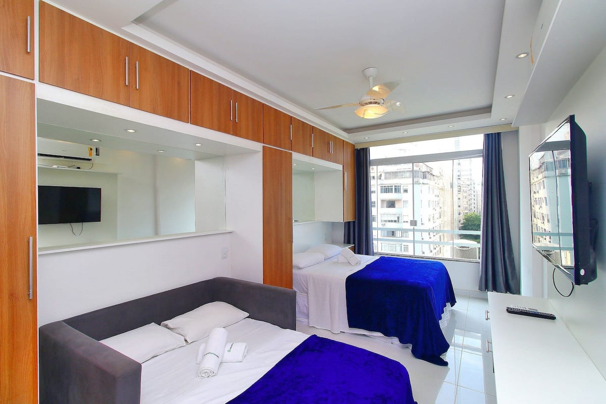 A hotel room with two beds, a desk, a TV, and a large window with a city view.