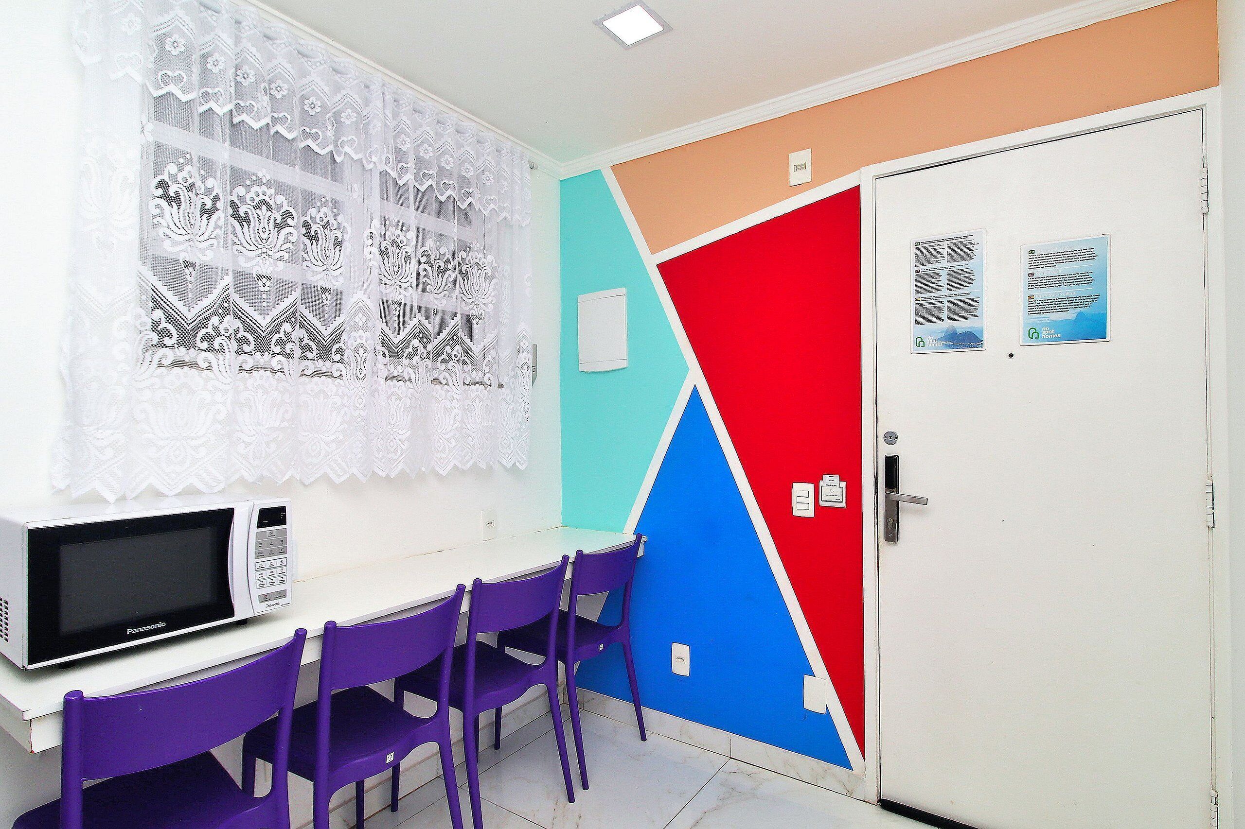 A room with a white table, purple chairs, a microwave, a window with lace curtains, and a door with informational posters.