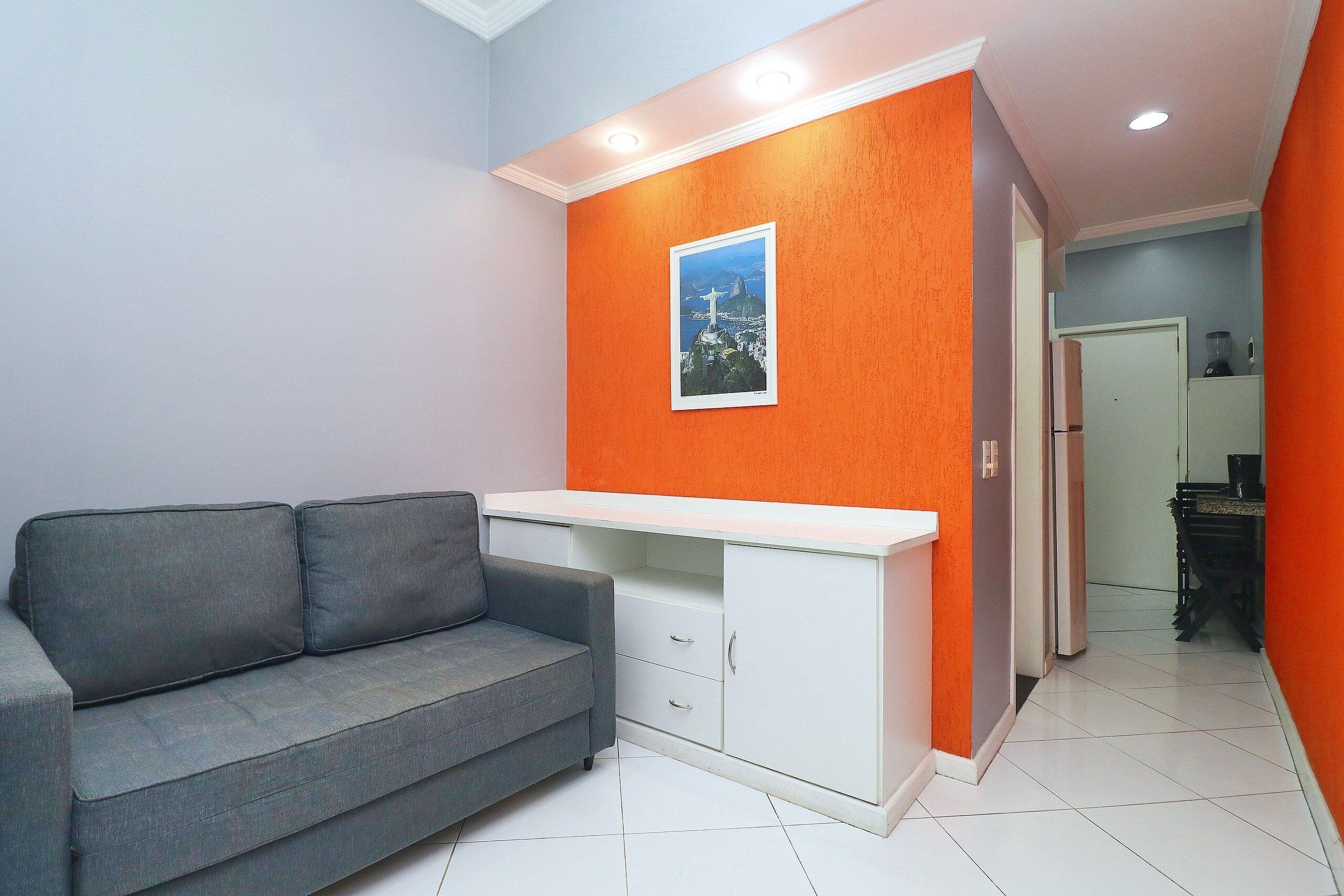 A compact living space with a grey sofa, a white desk, and an orange accent wall.