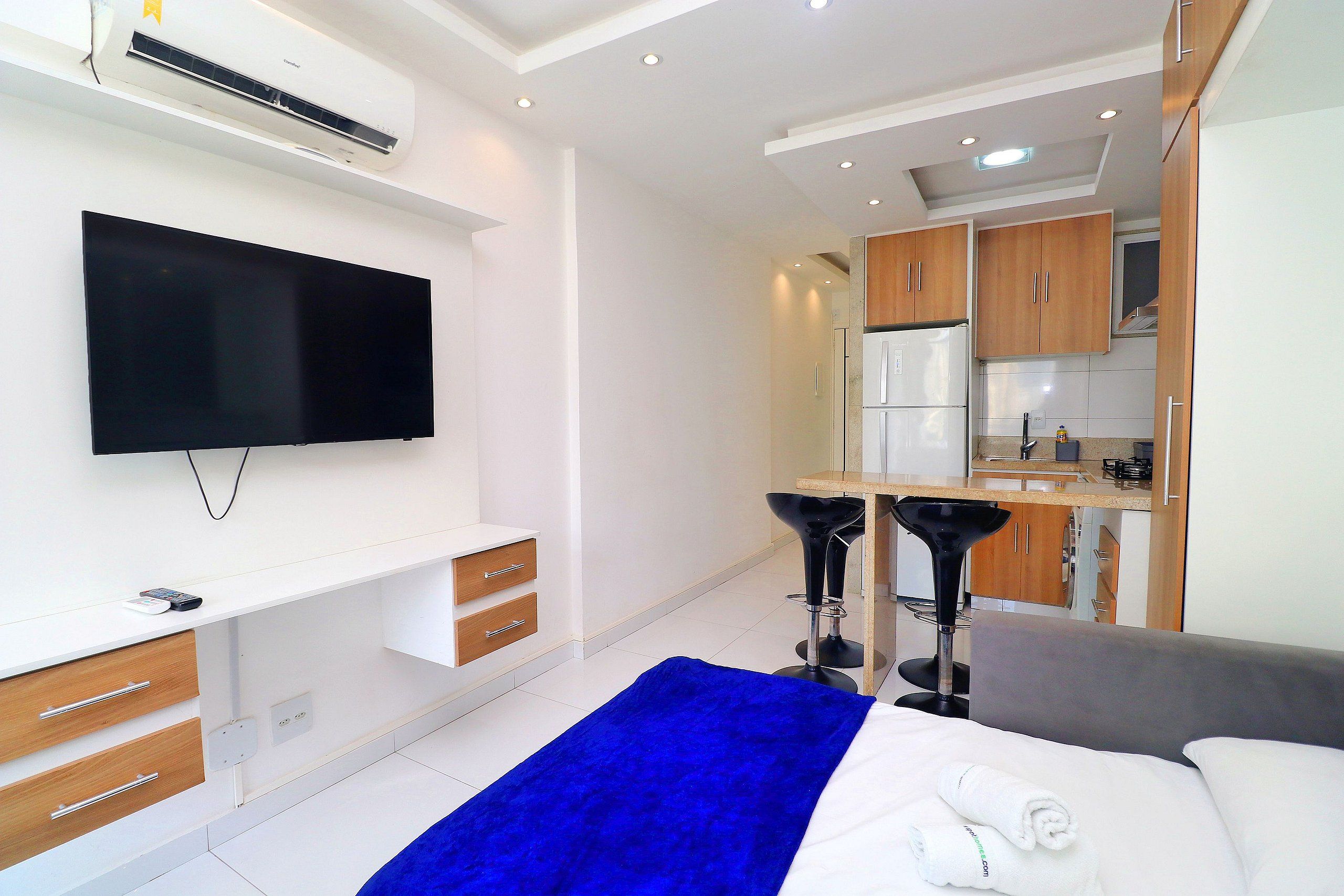 A modern hotel room with a flat-screen TV, a bed with a blue blanket, a grey sofa, a small kitchen area, and wooden cabinets.