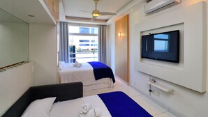 Quarto de hotel com duas camas, ventilador de teto, ar condicionado, televisão e janela com vista para prédios.