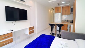 Quarto de hotel moderno com televisão de tela plana, cama com manta azul, sofá cinzento, pequena área de cozinha e armários de madeira.