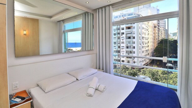 Quarto de hotel com cama grande, espelho e vista da cidade através de amplas janelas.