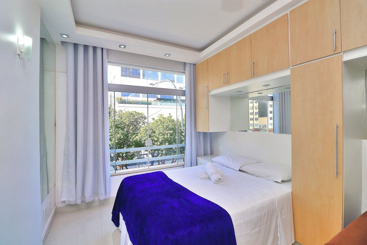 A hotel room with a large bed, a window with curtains, a wooden wardrobe, and a mirror above the bed.