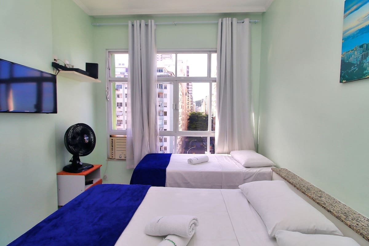 A hotel room with two beds, a TV, a fan, and a window with a city view.