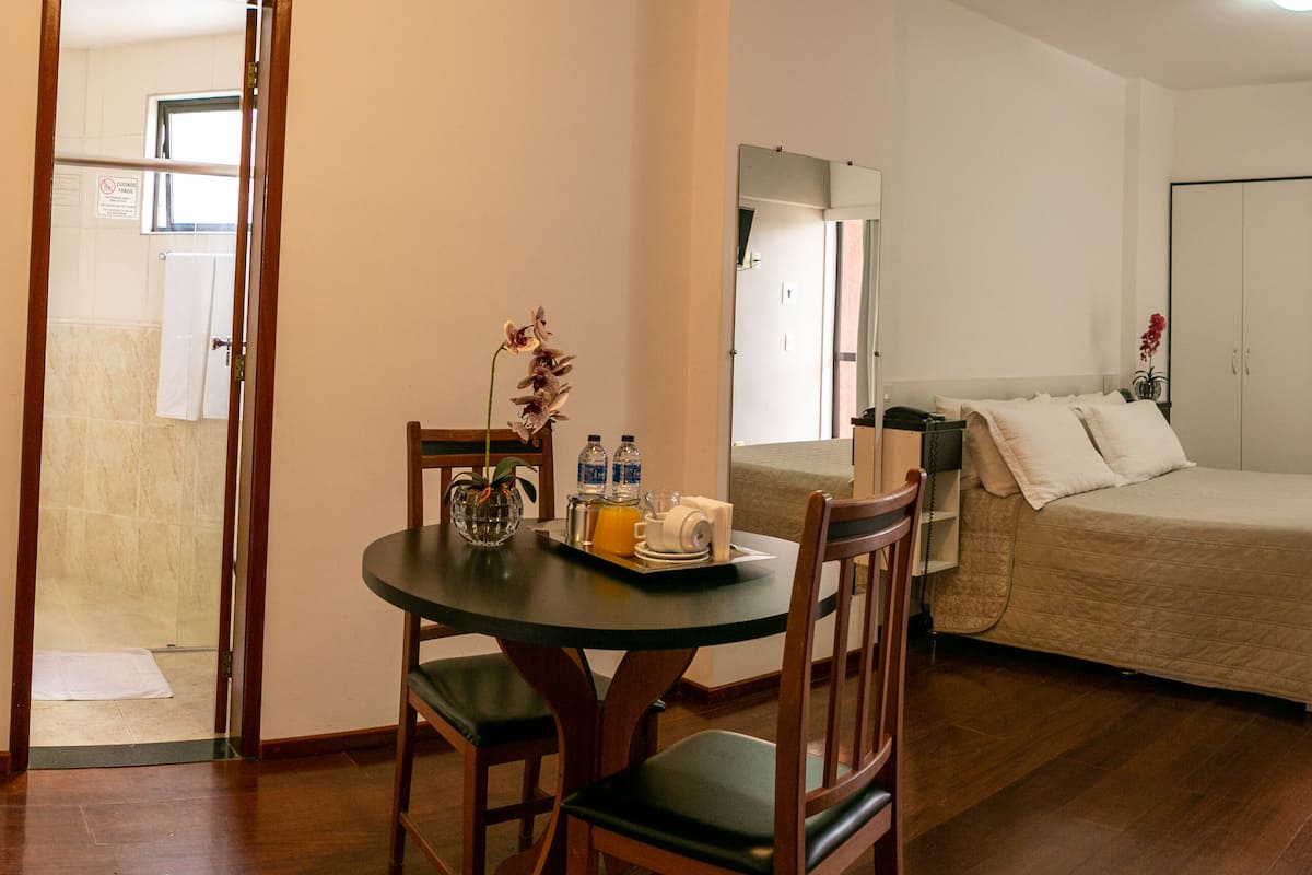 Una habitación de hotel con cama, una mesa con sillas, una mesita con un florero y botellas, y una puerta que da a un baño.