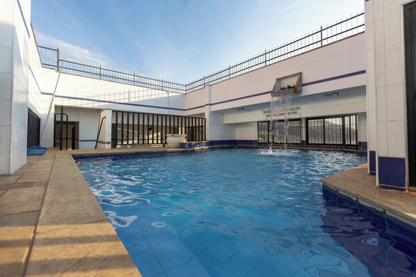 A swimming pool with a water fountain, surrounded by a building with large windows and a metal railing.