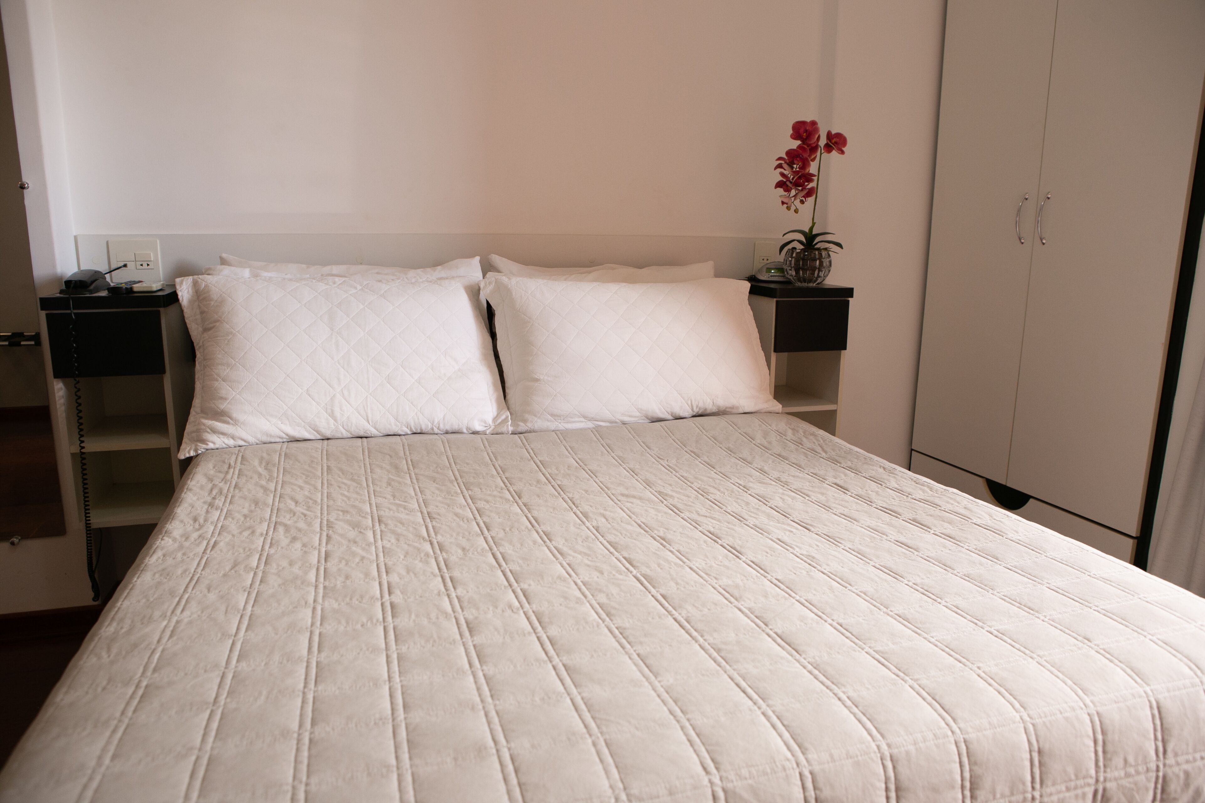A neatly made bed with white bedding, a bedside table with a lamp, and a vase with flowers on the nightstand.