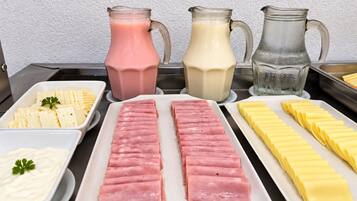 Mesa de buffet com diversos alimentos, incluindo presunto fatiado, queijo e um jarro de bebidas.