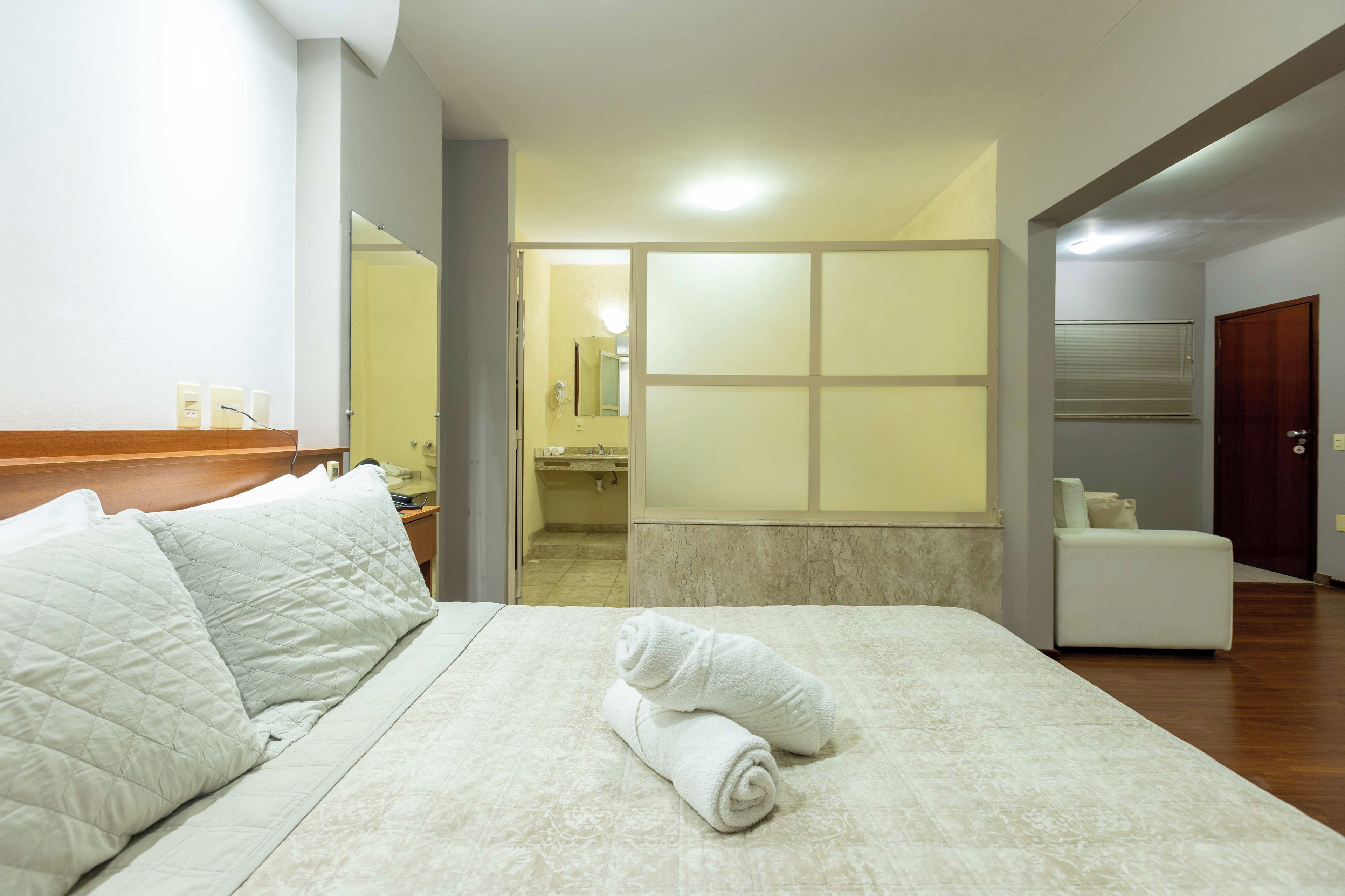 A hotel room with a bed, a bathroom visible through a glass door, and a seating area with a chair.