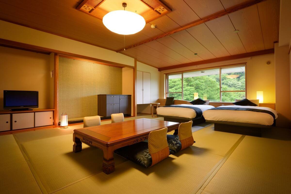 A traditional Japanese-style room with tatami flooring, a low wooden table, a bed with a view of greenery, and a television on a cabinet.