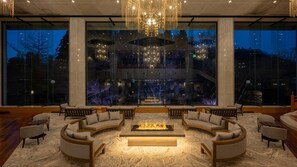 A modern lounge area with a central fire feature, comfortable seating, and large windows offering a view of the cityscape at night.