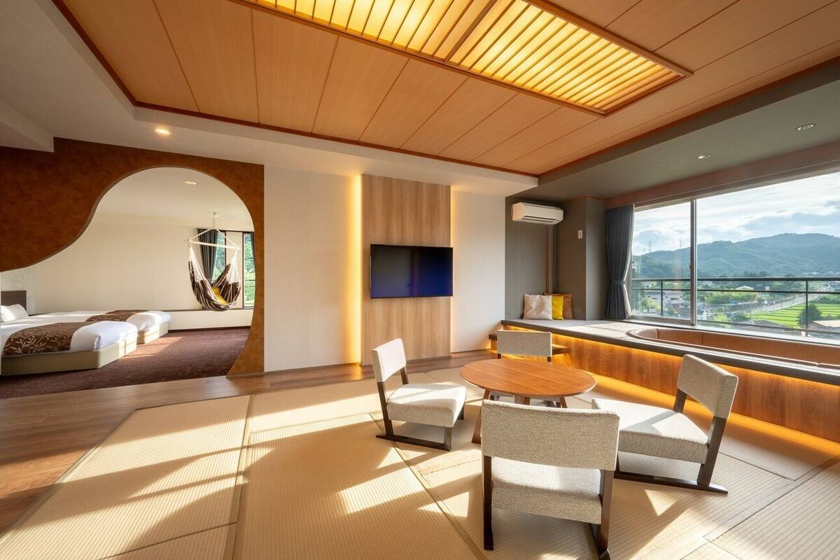 A modern hotel room with a large bed, a TV, a seating area with chairs, and a view of the mountains.
