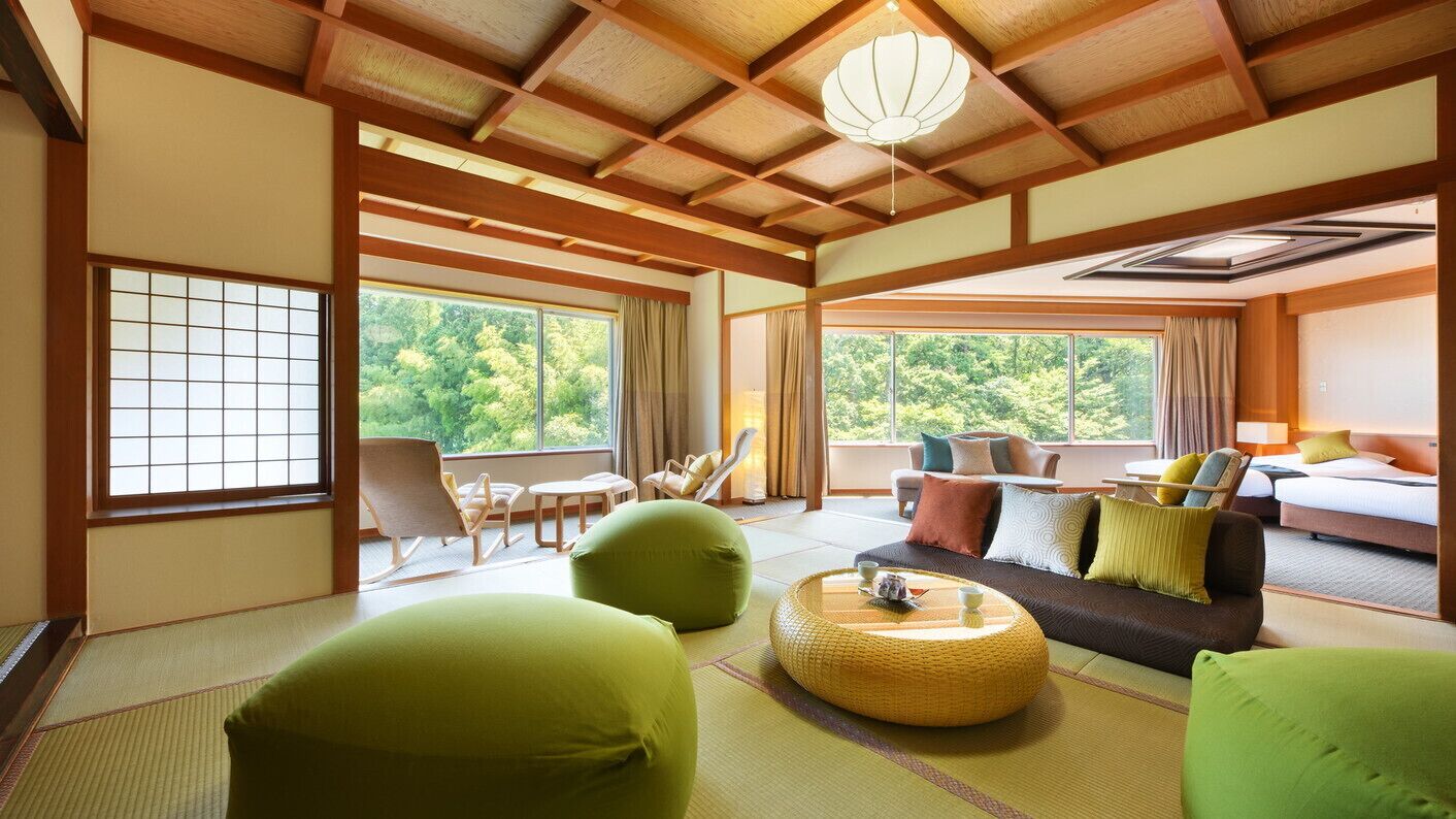 Ein traditioneller japanischer Raum mit Tatami-Böden, niedriger Decke mit Holzbalken, einer Couch mit Kissen, einem kleinen Tisch mit Teeservice und großen Fenstern mit Blick auf Grünflächen.
