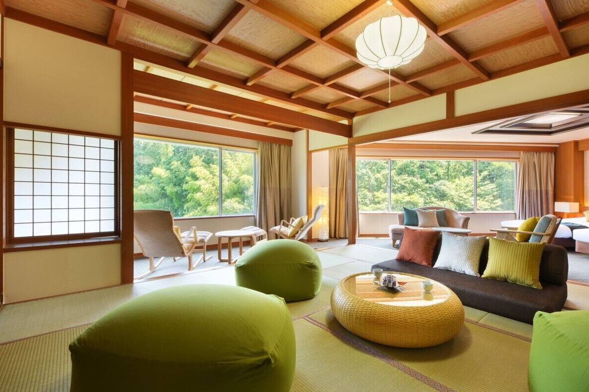 A traditional Japanese-style room with tatami flooring, a low ceiling with wooden beams, a sofa with cushions, a small table with a tea set, and large windows offering a view of greenery.