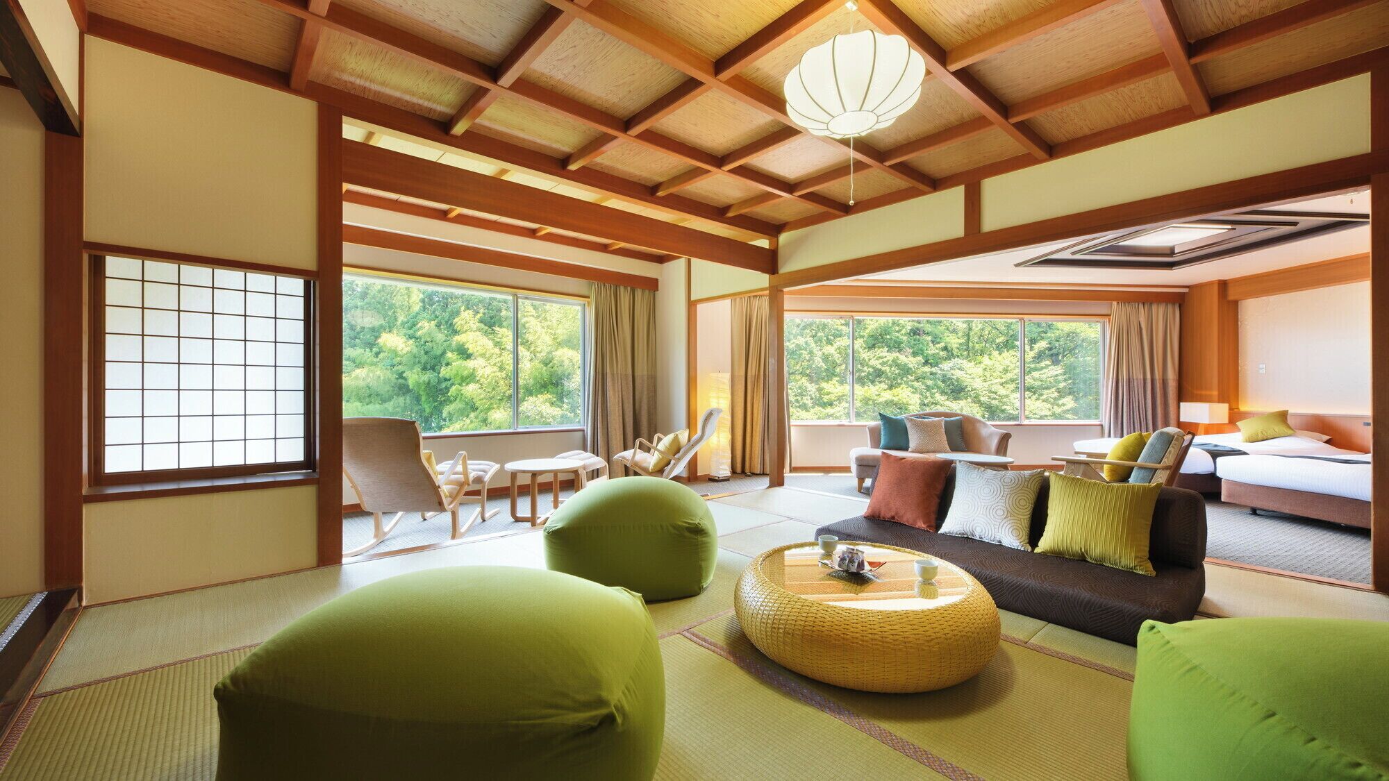 Ein traditioneller japanischer Raum mit Tatami-Böden, niedriger Decke mit Holzbalken, einer Couch mit Kissen, einem Esstisch und Blick ins Grüne.