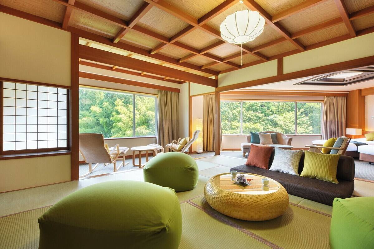 A traditional Japanese-style room with tatami flooring, a low ceiling with wooden beams, a sofa with cushions, a dining table, and a view of greenery outside.