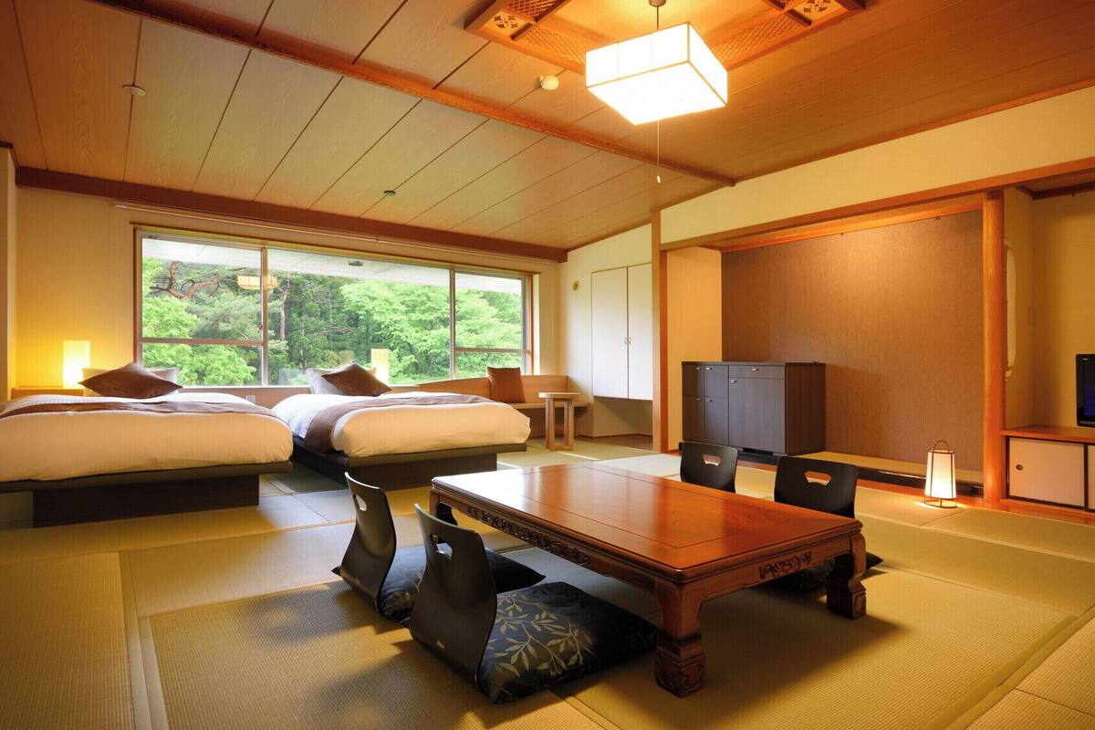 A traditional Japanese-style room with tatami flooring, a low wooden table, two beds, a television, and a view of greenery outside.