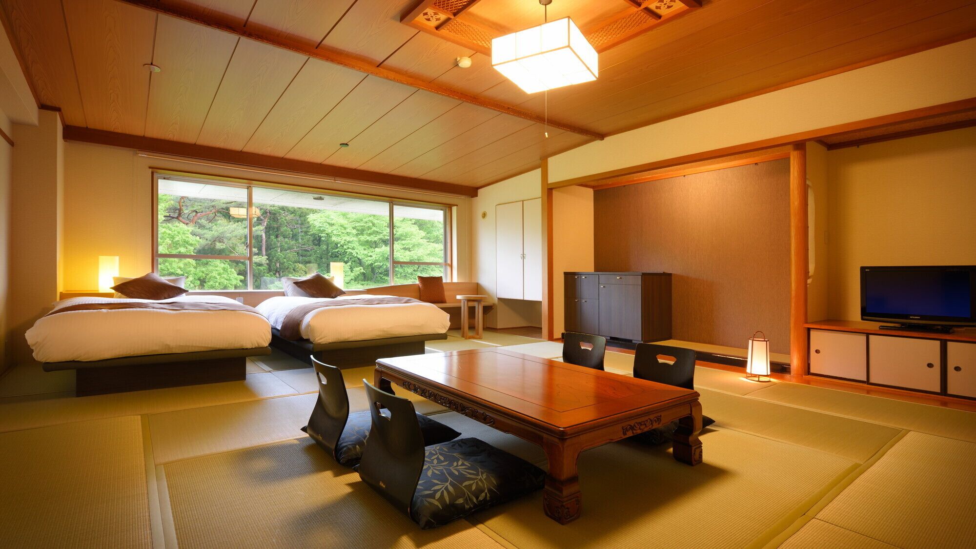 Ein traditioneller japanischer Raum mit Tatami-Böden, einem Holztisch, zwei Betten, einem Fernseher und einem großen Fenster mit Blick auf Grünflächen.