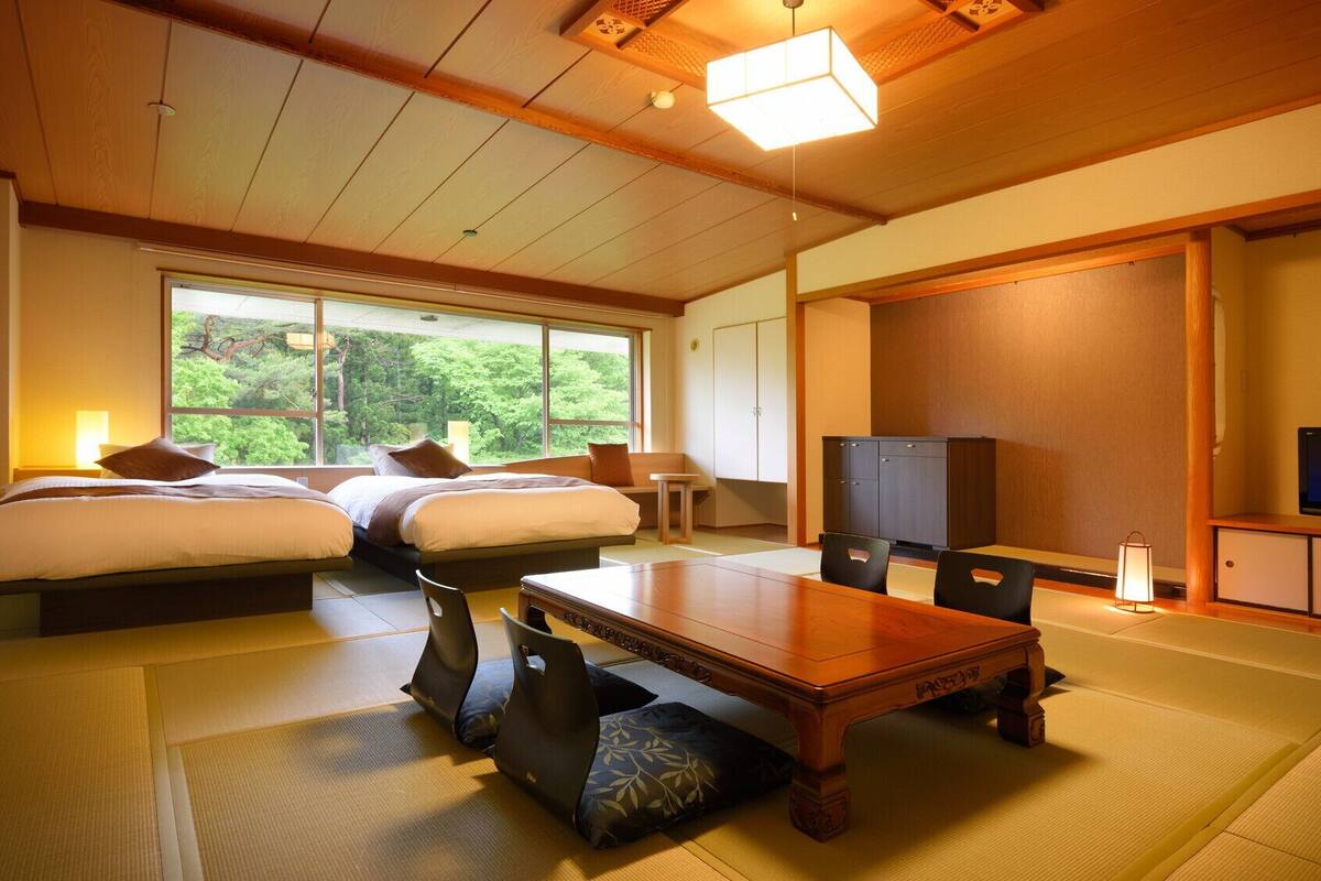 A traditional Japanese-style room with tatami flooring, a wooden dining table, two beds, a television, and a large window offering a view of greenery.