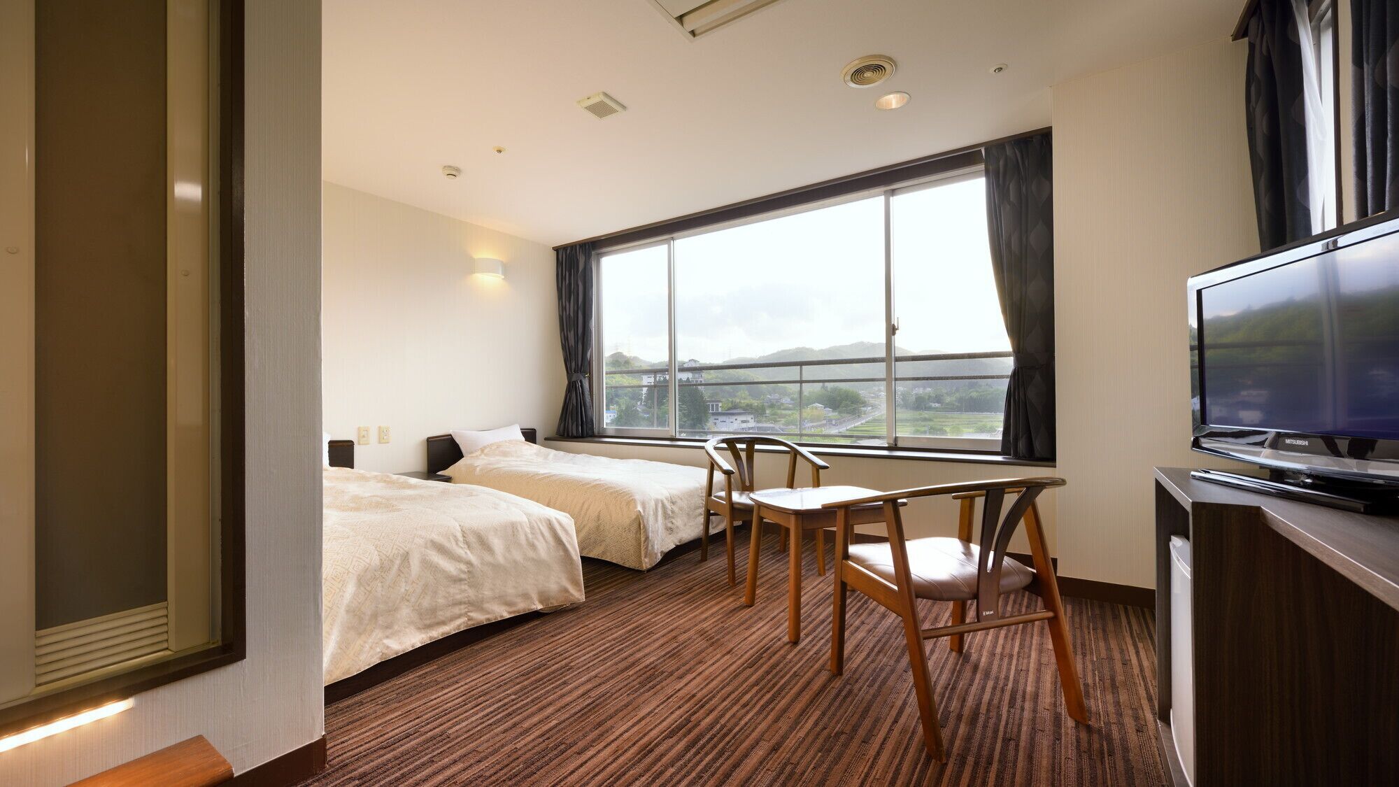 A hotel room with two beds, a TV, a desk, and a large window.