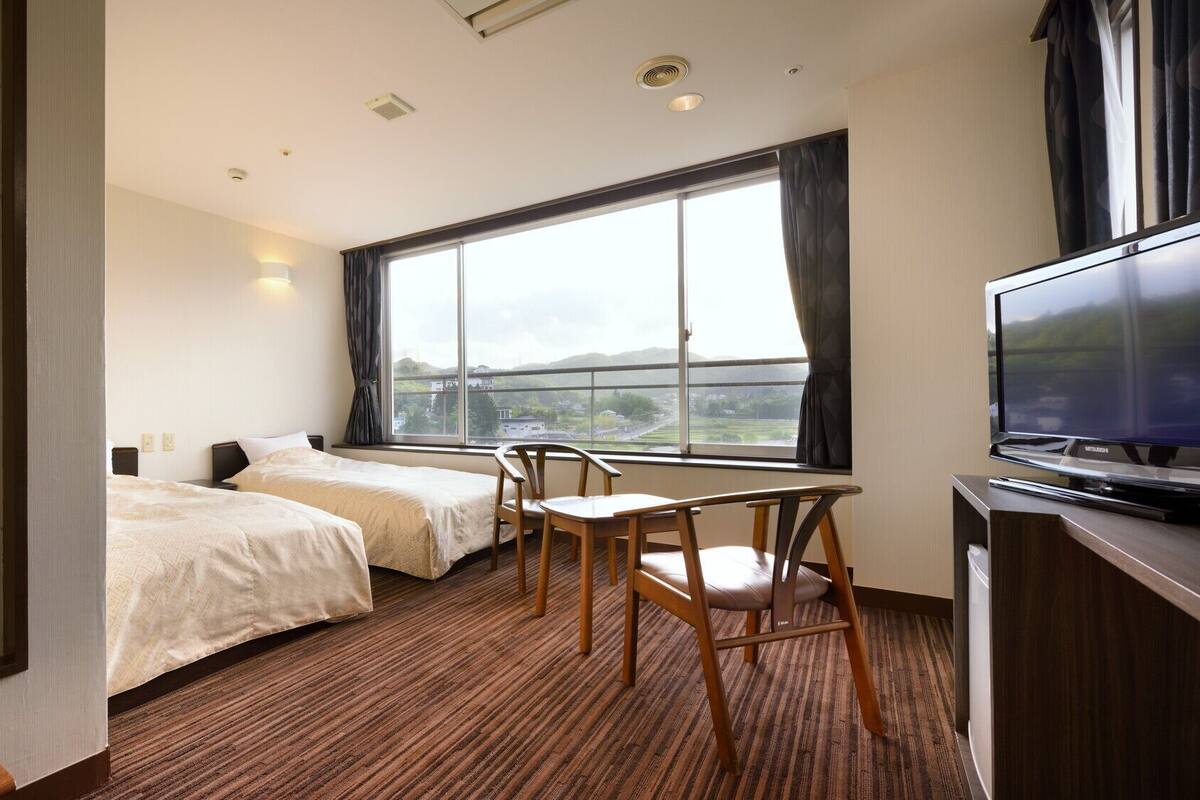 A hotel room with two beds, a TV, a desk, and a large window.