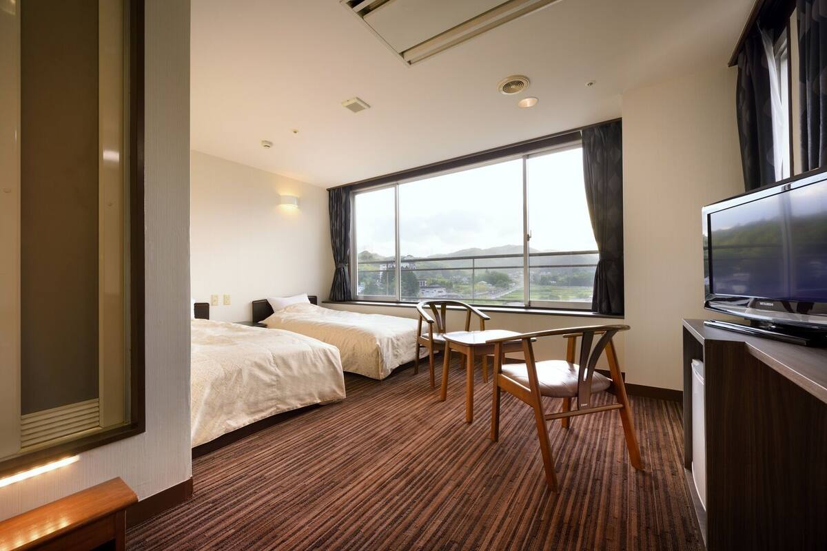 A hotel room with two beds, a TV, a desk with chairs, and a large window with a view.