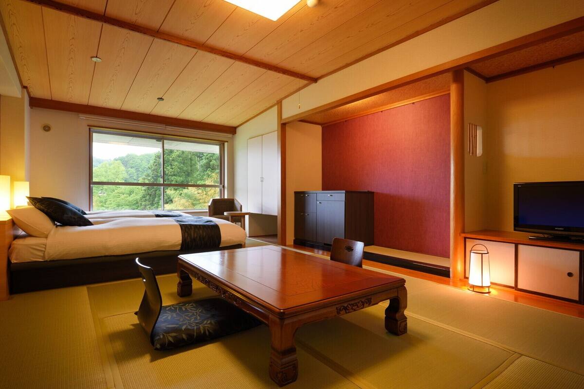 A hotel room with a bed, a television, a wooden table, and a view of greenery outside.