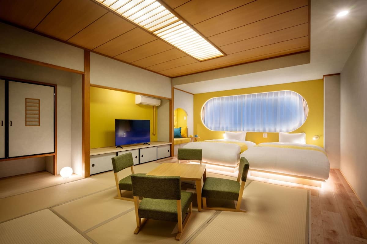 A modern hotel room with tatami flooring, a bed with a large window, a TV, and a seating area with chairs.