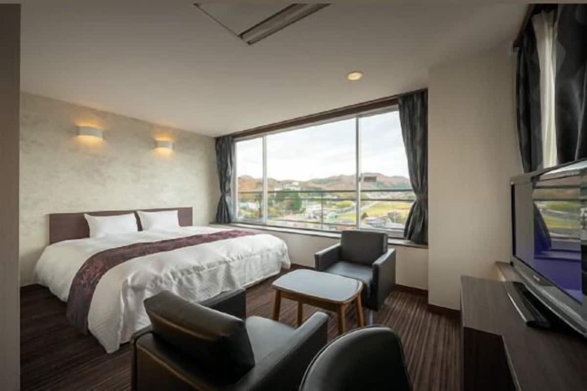 A hotel room with a bed, a TV, a desk, a chair, and a large window with a view.