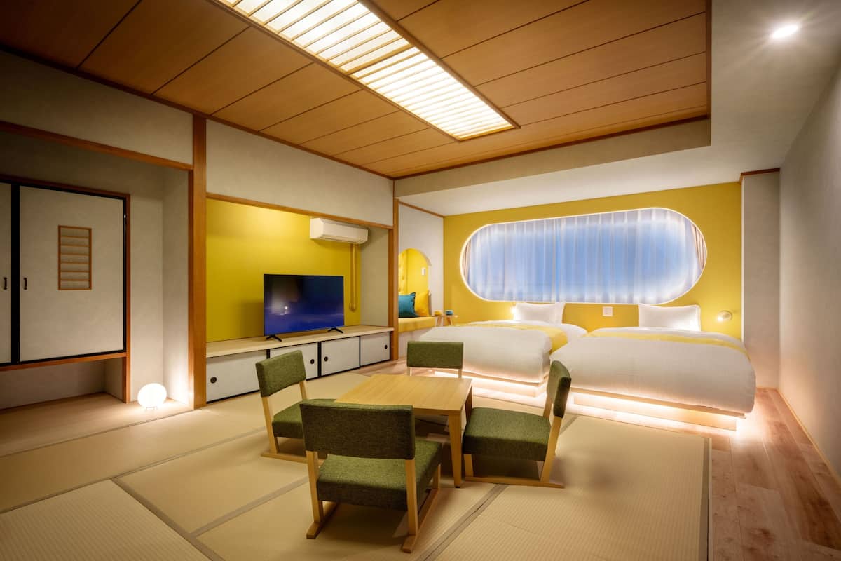 A modern hotel room with a bed, a TV, a dining area with chairs, and a traditional tatami mat floor.