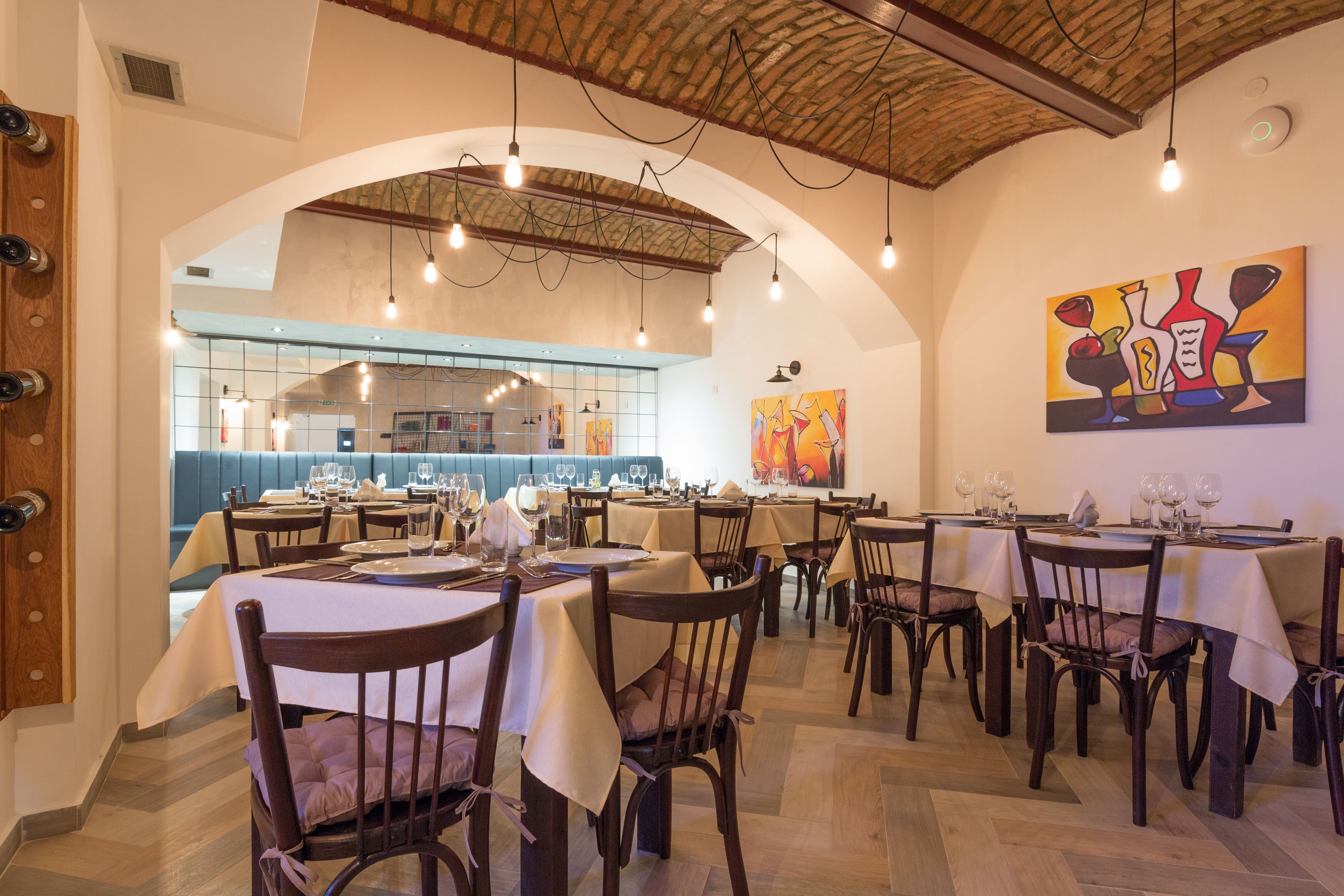 A restaurant with multiple tables and chairs, a kitchen in the background, and a large painting on the wall.