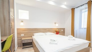A hotel room with a wooden headboard, a bed with white linens, a desk with a chair, and a window with curtains.