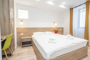 Ein Hotelzimmer mit Holz-Kopfteil, einem Bett mit weißer Bettwäsche, einem Schreibtisch mit Stuhl und einem Fenster mit Vorhängen.