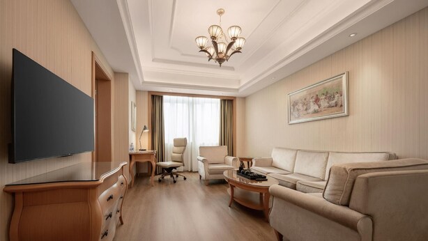 A hotel room with a flat-screen TV, a sofa, a coffee table, a desk, and a chandelier. - Vienna Hotel Foshan Nanhai Avenue Branch (Foshan)